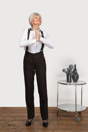 Front view of an old pleased lady in office clothing holding hands together while looking up