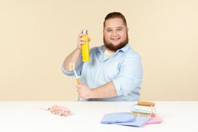 Contented young overweight man sitting at the table and holding cleaning equipment