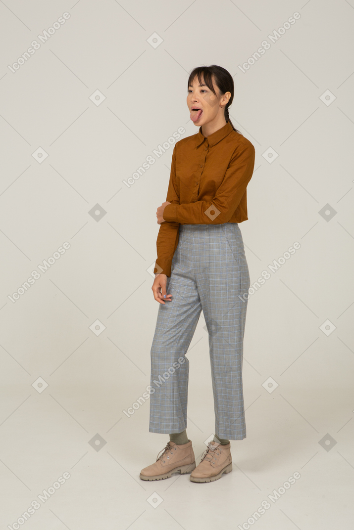 Vista lateral de uma jovem mulher asiática de calça e blusa tocando o braço e mostrando a língua