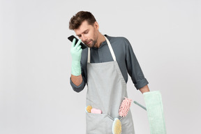 Young househusband holding mop and in another hand phone close to his mouth