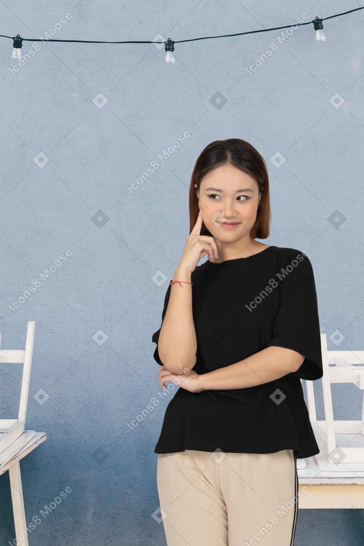 Застенчивая женщина в черной футболке стоит с указательным пальцем на щеку
