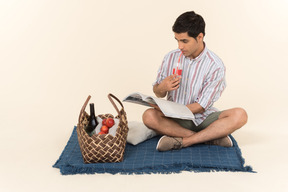 Young caucasian guy sitting on blanket, reading magazine and drinking a coke