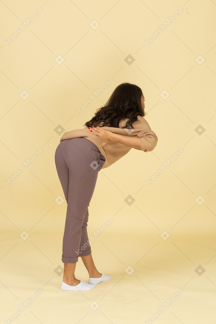 Vista posterior de tres cuartos de una mujer joven de piel oscura que se inclina hacia adelante mientras pone las manos en las caderas