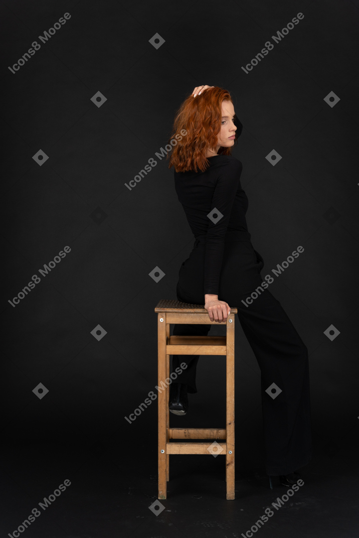 Una vista lateral trasera de la hermosa joven sentada en la silla alta de madera y ajustando su cabello