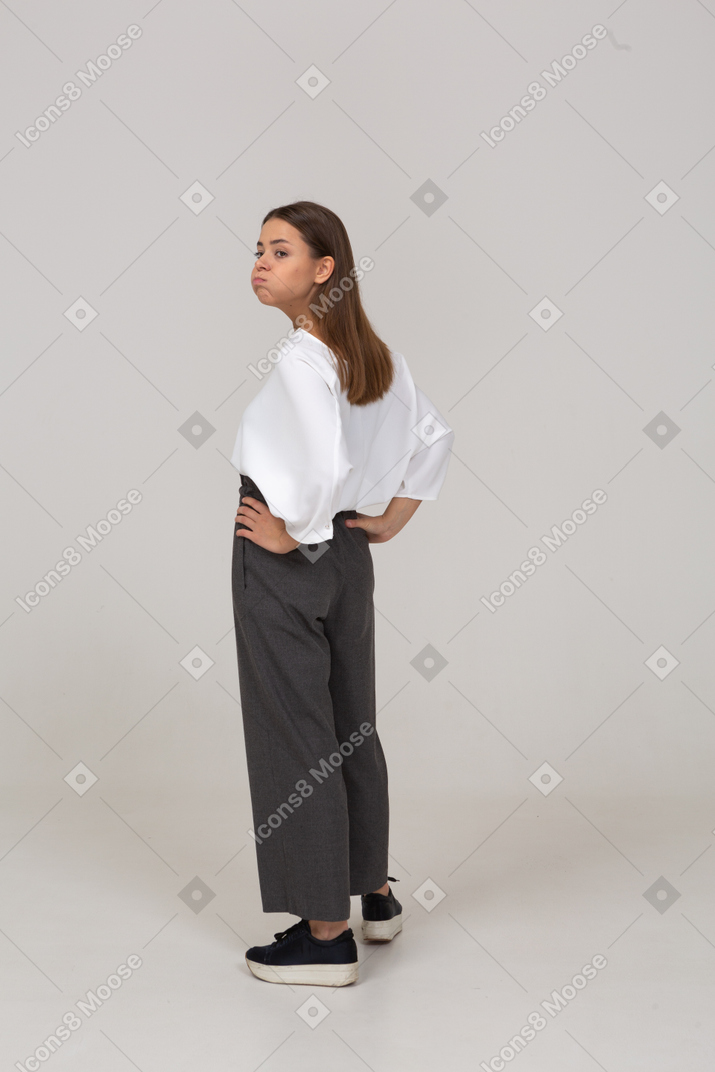 Dreiviertel-rückansicht einer jungen dame in bürokleidung, die wangen bläst und die hände auf die hüften legt