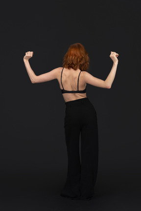 A back side view of the cute young woman dancing on the black background holding her fists up