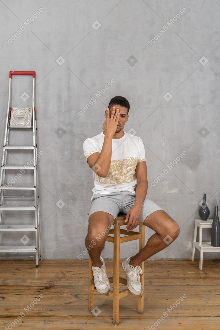 Junger mann sitzt auf einem stuhl und bedeckt ein auge mit der hand