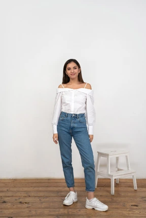 A young woman standing next to a white chair in front of a white wall and smiling