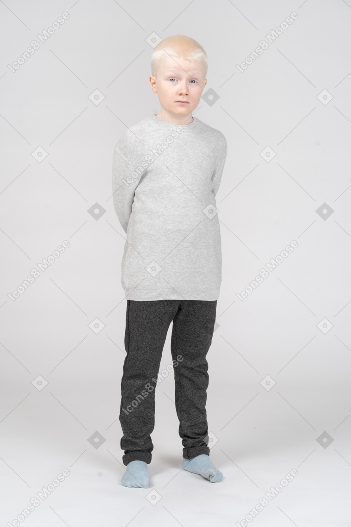 A blonde little boy in casual clothes standing and hiding hands behind