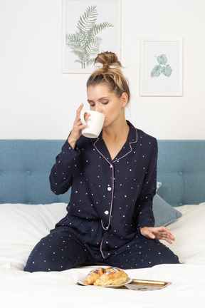 Front view of a young lady in pajamas drinking coffee in bed