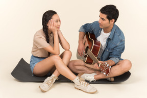 Young caucasian guy playing guitar sitting near asian woman on karimat