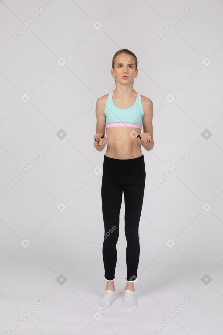 Vista frontal de una perpleja jovencita en ropa deportiva levantando las manos y mirando hacia arriba