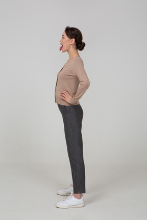 Vista laterale di una giovane donna in pullover e pantaloni che mette le mani sui fianchi e mostra la lingua