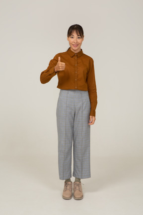 Vista frontal de uma jovem mulher asiática de calça e blusa aparecendo o polegar