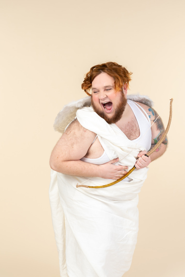 Laughing young  man dressed as a cupid holding bow and arrow and pointing a