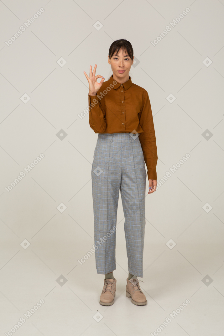 Vista frontal de uma jovem mulher asiática de calça e blusa, mostrando um gesto de ok