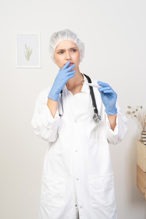 Front view of a worried young female doctor with stethoscope holding thermometer