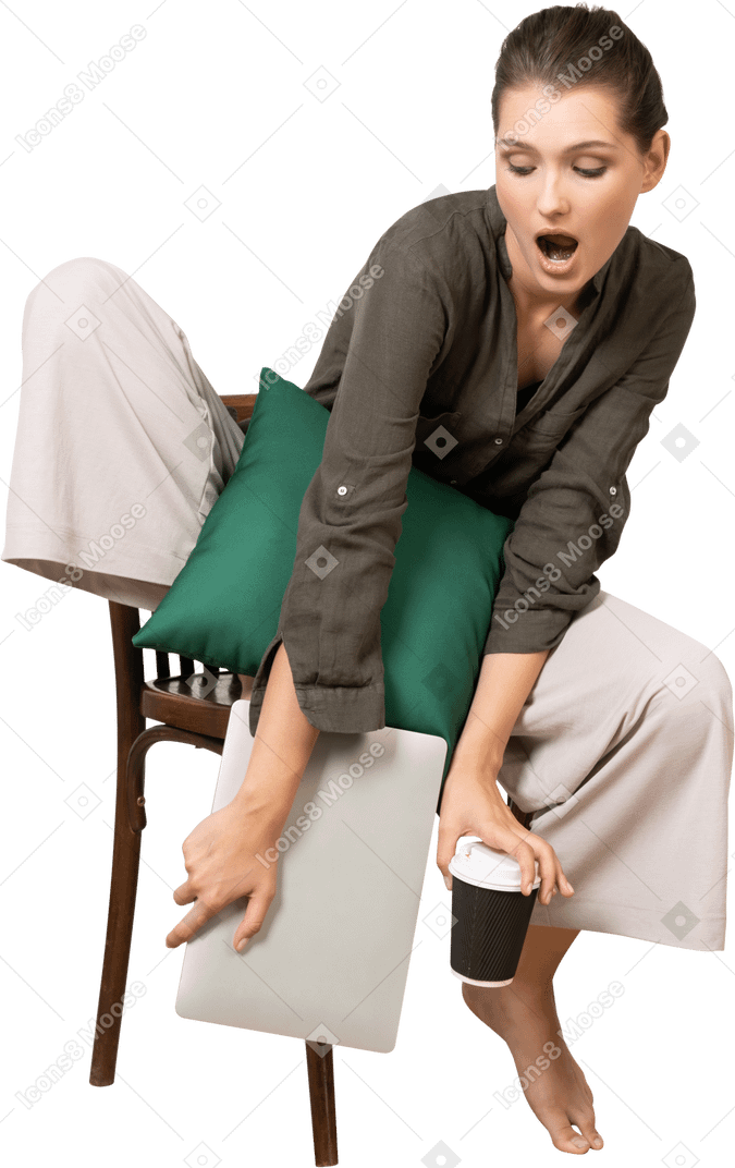 Вид спереди потрясенной молодой женщины, сидящей на стуле, держащей ноутбук и трогательной чашкой кофе