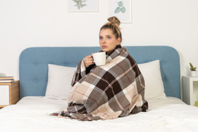Vista lateral de una joven enferma envuelta en una manta marcada en la cama sosteniendo una taza de té