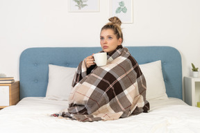 Side view of an ill young lady wrapped in checked blanket in bed holding a cup of tea