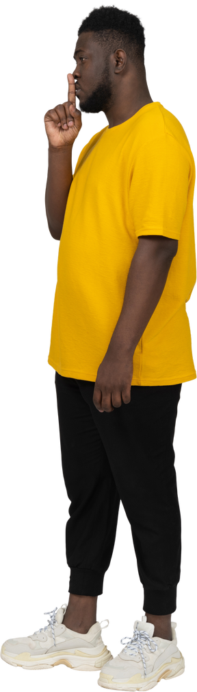 Vista di tre quarti di un giovane uomo dalla pelle scura in maglietta gialla che mostra gesto di silenzio