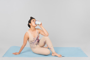 Young indian gymnast sitting on yoga mat and drinking coffee