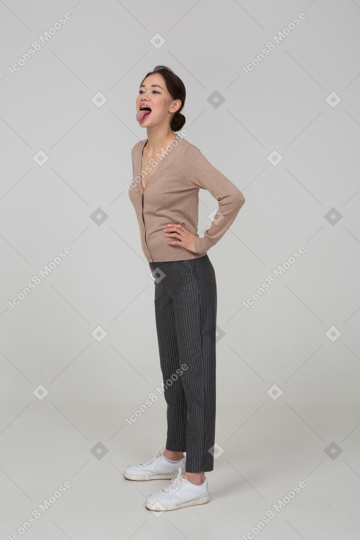 Vista de três quartos de uma jovem senhora de pulôver e calça, colocando as mãos nos quadris e mostrando a língua