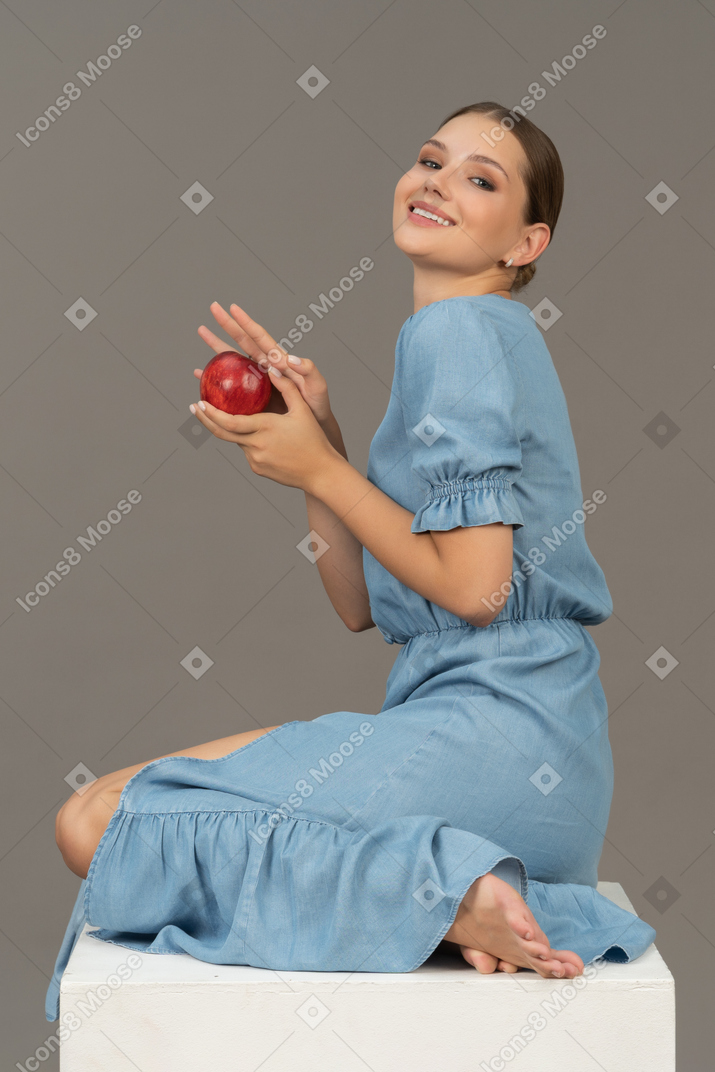 Side view of cheerful young woman with apple sitting on cube and looking at camera