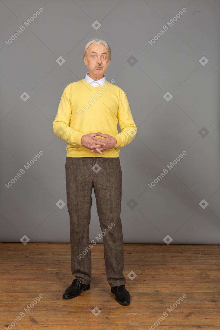 Вид спереди надутого старика в желтом свитере, смотрящего в камеру и держащего руки вместе