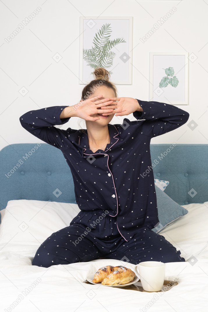 Front view of a young lady in pajamas hiding face before having breakfast in bed
