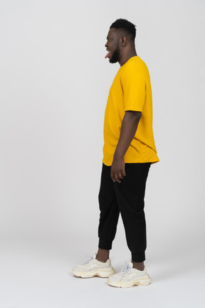 Side view of a young dark-skinned man in yellow t-shirt standing still & showing tongue