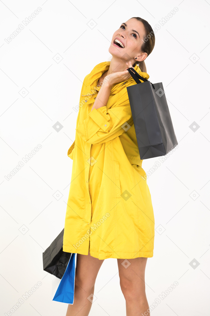 Femme souriante dans un manteau jaune tenant un sac à provisions sur une épaule et regardant quelque part