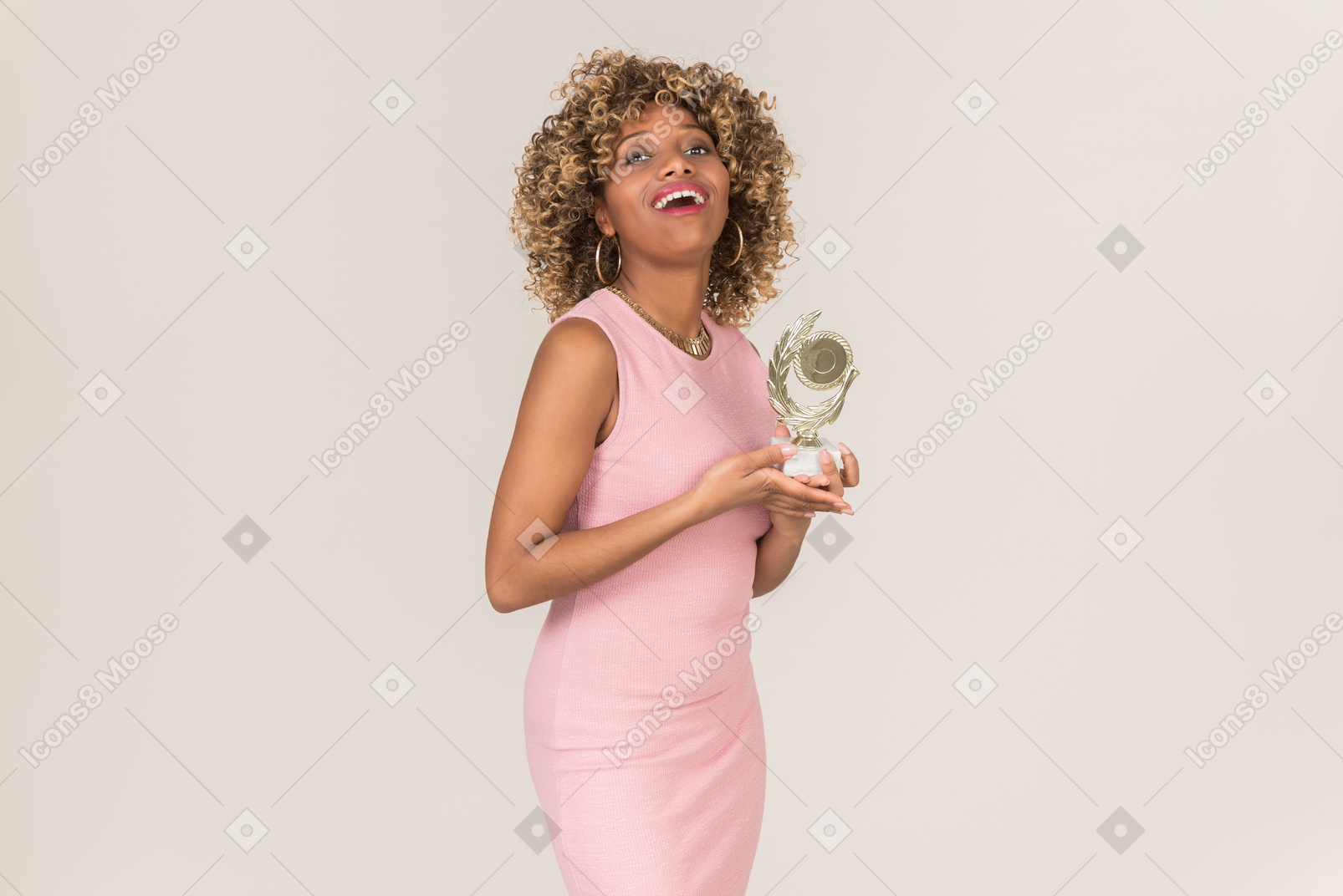 A young black fluffy-haired woman in a pastel pink cocktail dress, standing with a prize in her hands, against a plain grey background