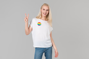 Beau jeune homme aux longs cheveux blonds, debout sur un fond gris, vêtu d'un jean bleu et d'un t-shirt blanc avec écusson lgbt