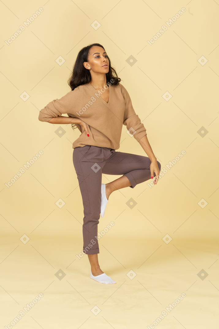 Вид в три четверти темнокожей молодой женщины, которая кладет руку на бедро, поднимая ногу