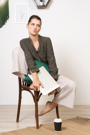 Front view of a pouting young woman sitting on a chair and holding her laptop