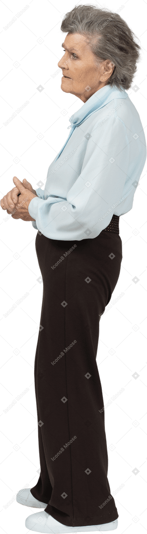 Vista laterale di una donna anziana seria vestita in camicetta e pantaloni che tengono le mani insieme