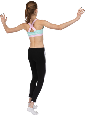 Vista posterior de tres cuartos de una jovencita en ropa deportiva balanceándose de puntillas mientras levanta las manos
