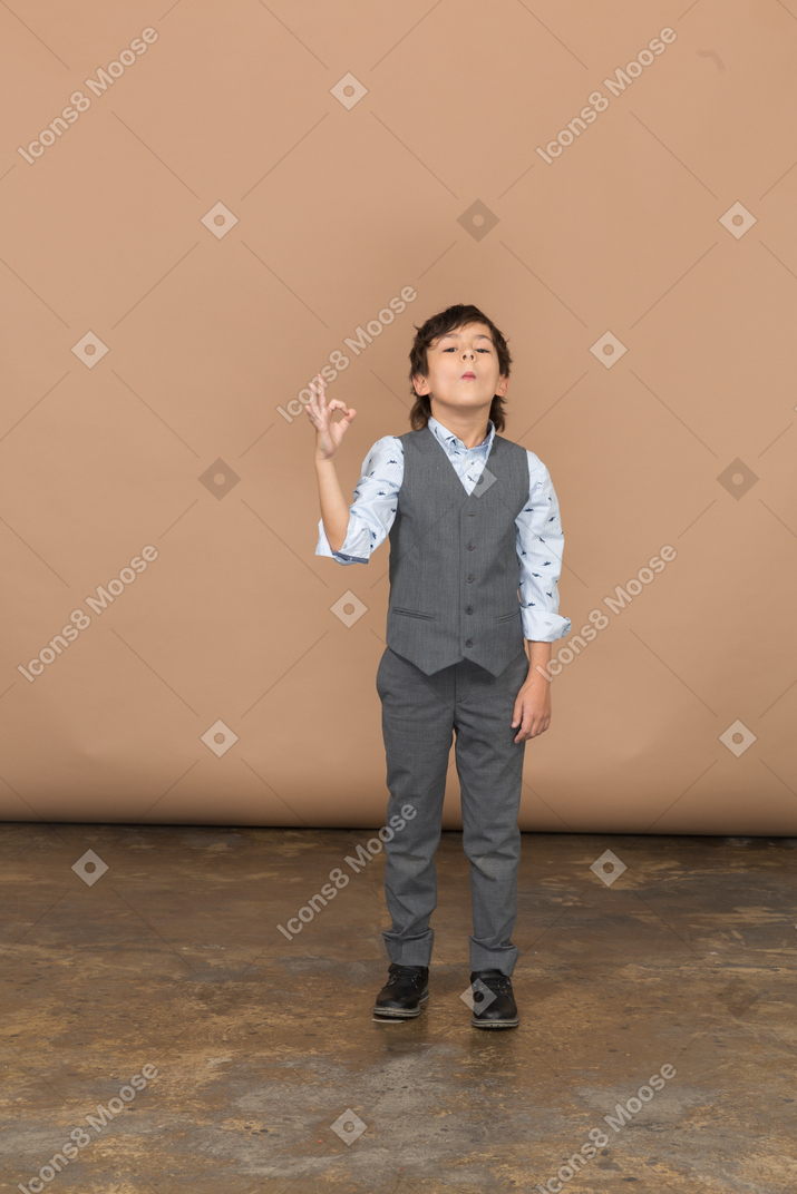Вид спереди на симпатичного мальчика в сером костюме, показывающего знак ок