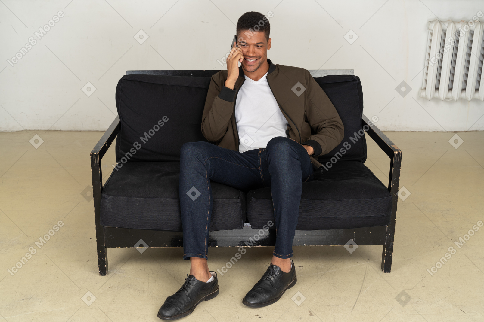 Vista frontal de um jovem sorridente, sentado em um sofá e falando ao telefone