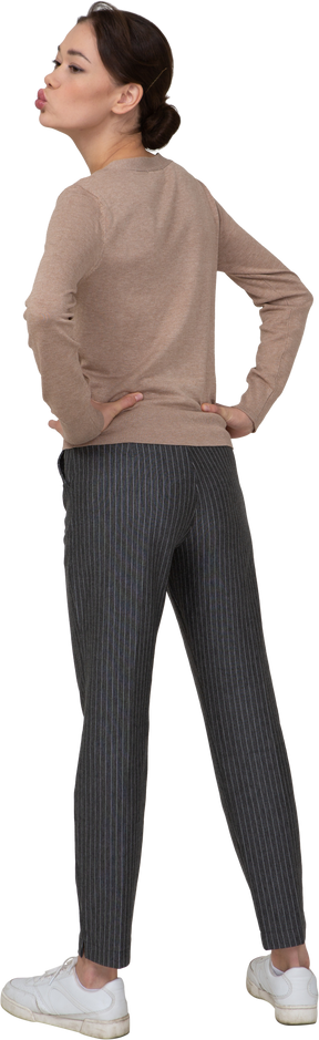 Vista posterior de una joven en suéter y pantalones enviando un beso al aire y poniendo las manos en las caderas