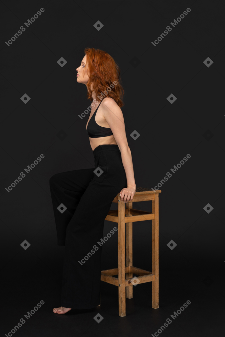 A side view of the beautiful woman dressed in black pants and bra, sitting on the wooden chair and looking to the left