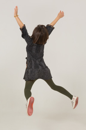 Back view of a jumping little girl in dress outspreading hands and legs