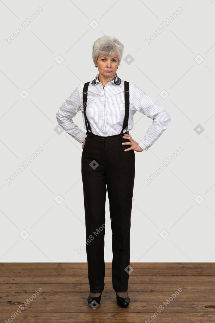 A figura intera di una vecchia femmina perplessa in bretelle che mette le mani sui fianchi