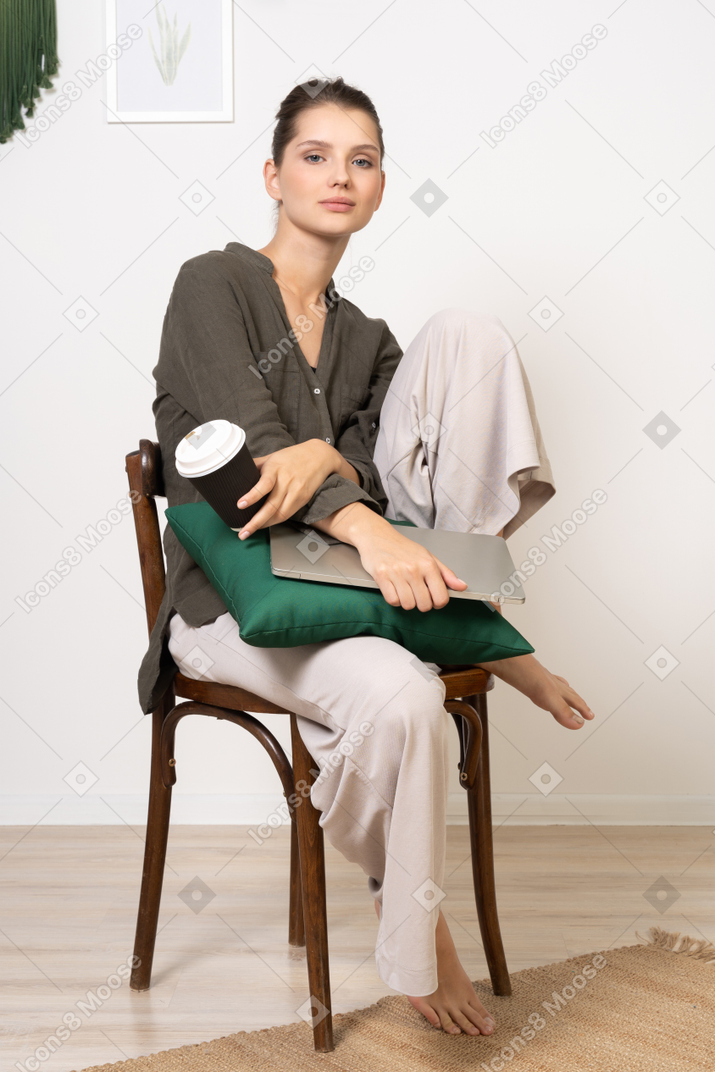 Three-quarter view of a young woman sitting on a chair and holding her laptop & touching coffee cup