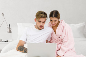 A man and woman sitting on a bed looking at a laptop