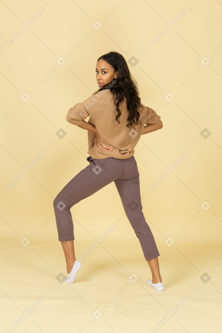 Three-quarter back view of a dark-skinned young female putting hand on hip while bending knee
