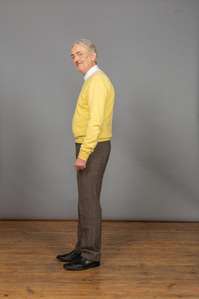 Вид сбоку на пожилого веселого человека в желтом пуловере, наклонившегося и смотрящего в камеру, морщась