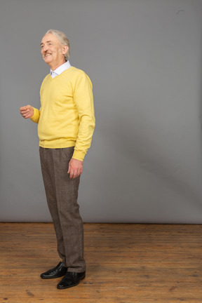 Vista de três quartos de um velho gesticulando vestindo um pulôver amarelo e olhando para o lado enquanto sorri
