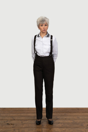 Front view of an old funny female in office clothes grimacing with her hands behind back